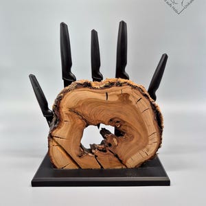 Könnte beinhalten: Ein Messerblock aus Holz in Form einer Hand, der fünf schwarze Messer hält. Der Block besteht aus einem Querschnitt eines Baumstamms mit natürlicher Kante und einem Loch in der Mitte. Der Sockel ist schwarz.