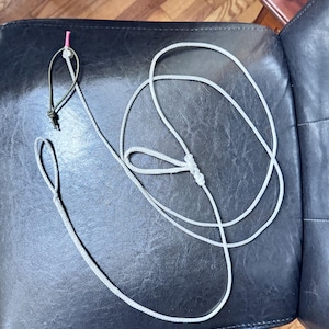 May include: A gray rope dog leash with a loop handle and a loop at the end. The leash has a green and black loop with a pink connector. The leash is lying on a black leather surface.