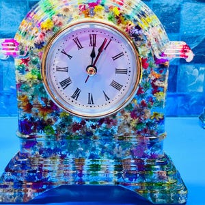 May include: A decorative clock with a white face, gold trim, and black Roman numerals. The clock is encased in a clear resin filled with colorful flowers. The clock's base and arched top are also made of the same resin.