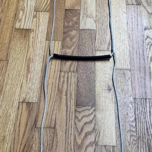 May include: A rope ladder with three black rungs and silver-gray rope sides. The ladder is designed for climbing and has metal loops at the top for hanging. The ladder is lying on a wooden floor.