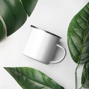 Pode incluir: Uma caneca de esmalte branco com borda e alça prateadas, sobre um fundo branco e rodeada por grandes folhas verdes. A caneca está vazia e pronta para uso.