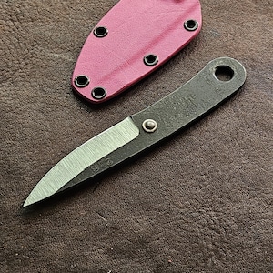 May include: A small, handmade knife with a dark, textured handle and a silver blade. The blade has the number "238" etched on it. A pink sheath with black eyelets is next to the knife. The knife and sheath are on a brown leather surface.