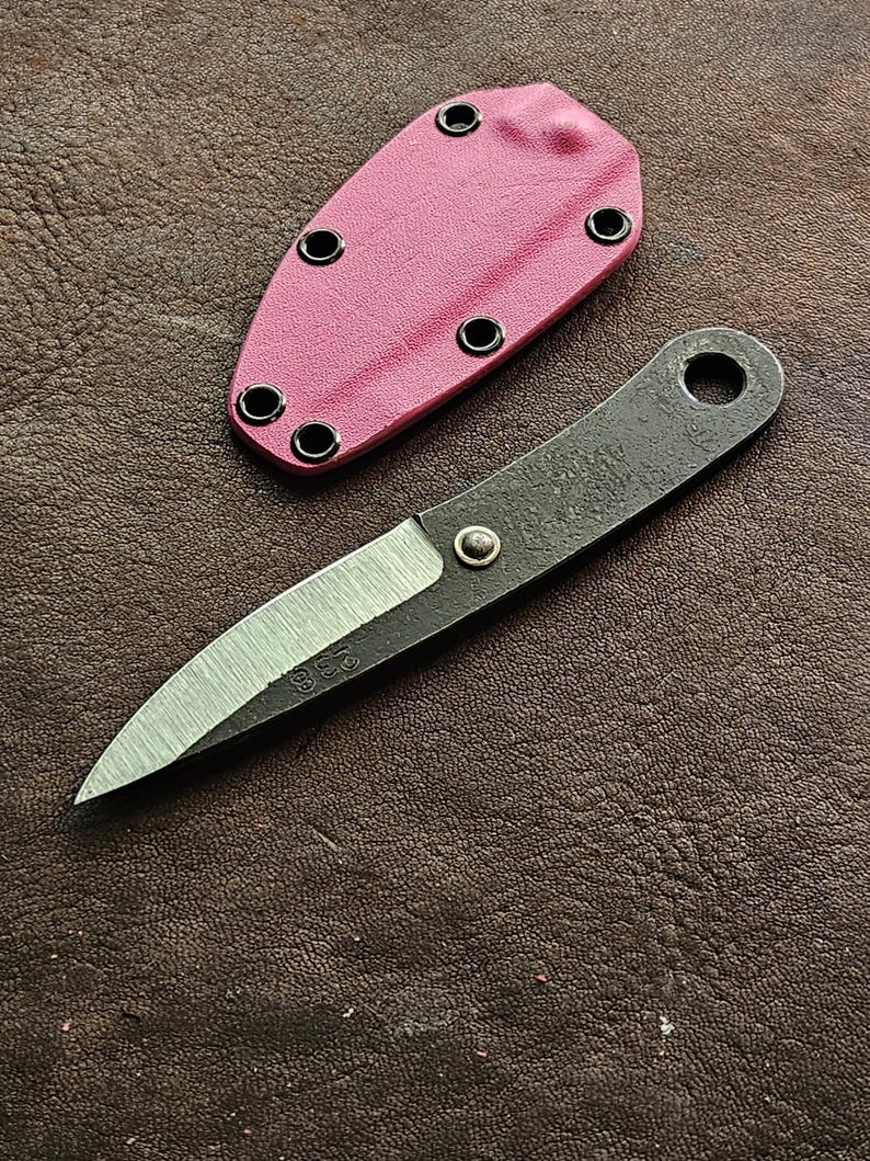 May include: A small, handmade knife with a dark, textured handle and a silver blade. The blade has the number "238" etched on it. A pink sheath with black eyelets is next to the knife. The knife and sheath are on a brown leather surface.