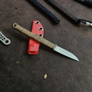 Op de afbeelding: Een mes met een vaststaand lemmet van zilverkleurig metaal en een handvat omwikkeld met beige koord. Het mes ligt op een felrode schede met drie zilverkleurige metalen oogjes aan de zijkant, met andere messen zichtbaar in de buurt.