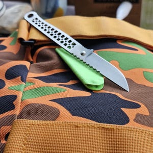 May include: A folding knife with a silver-toned metal handle featuring a perforated pattern and a serrated blade, resting on a bright lime green protective sheath placed atop a fabric bag with an orange, green, and black camouflage pattern.