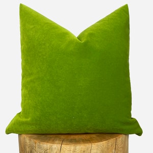 May include: A green velvet pillow with a square shape. The pillow is sitting on a wooden stump.