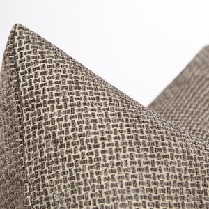 May include: Close-up of a brown and beige woven pillow cover with a textured pattern. The fabric has a subtle sheen and a slightly raised weave.