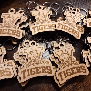 May include: A collection of wooden keychains featuring the text "TIGERS" in a bold font. Each keychain has a tiger graphic and the word "VALENCIA" above the text. The keychains are made of light-colored wood and have silver key rings.