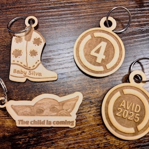 May include: Four wooden keychains with different designs. One keychain features a cowboy boot with the text "Baby Silva". Another keychain is round with the number "4" inside a circle. The third keychain is round with the text "AVID 2025" inside a circle. The fourth keychain features a cartoon image of a sleeping baby with the text "The child is coming".