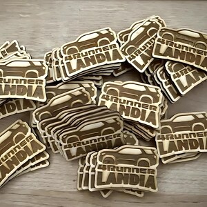 May include: A pile of wooden laser cut stickers with the text "4Runner Landia" on them. The stickers are brown and feature a silhouette of a car.