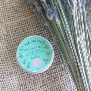 May include: A small, round, green tin with a white label that reads "THE MIN/ Wonder Balm LAVENDER ESSENTIAL OIL & APRICOT CARRIER OIL Handmade by 15ML". The tin is surrounded by lavender flowers.