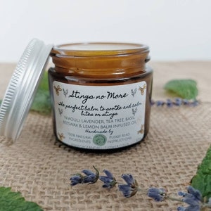 May include: A small amber glass jar with a silver lid, labelled "Stings no More" and containing a balm to soothe and calm bites or stings. The label lists ingredients including Niaouli Lavender, Tea Tree Basil, Beeswax, and Lemon Balm infused oil. The jar is sitting on a hessian surface with green leaves and purple flowers.