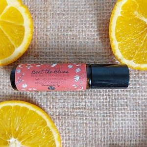 May include: A red and white rollerball bottle with the text "Beat the Blues" and a list of ingredients: bergamot, frankincense, patchouli, ginger, and apricot carrier oil. The bottle is on a brown hessian surface with orange slices.