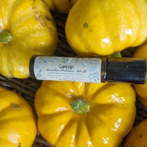 May include: A small glass bottle with a black roller ball top. The bottle is labelled "Samhain Connection Protection Rebirth" and lists the essential oils included: clary sage, mandarin, nutmeg, sandalwood, apricot, and jojoba. The bottle is sitting on a woven basket with yellow squash.