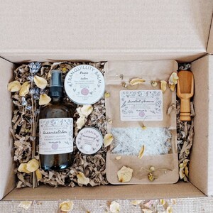 May include: A brown gift box containing self-care items. Inside are a spray bottle, bath salts, lip balm, and a balm. The box is filled with shredded paper and dried flowers. The products have labels with botanical illustrations and text.