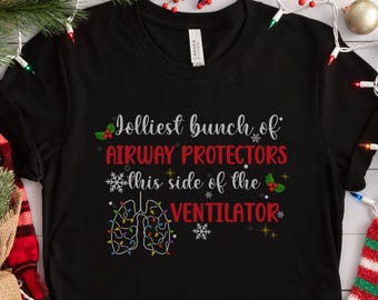 May include: Black t-shirt with the text "Jolliest bunch of AIRWAY PROTECTORS this side of the VENTILATOR." The design includes a lung graphic decorated with Christmas lights. Festive socks and holiday decorations are in the background.