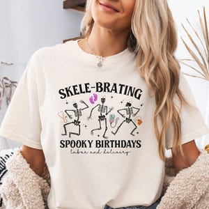 May include: Ivory-colored t-shirt with the text "SKELE-BRATING SPOOKY BIRTHDAYS labor and delivery." The shirt features a graphic of three dancing skeletons with medical-themed accessories. The shirt is paired with denim shorts.
