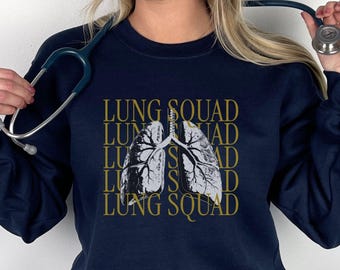 May include: A navy blue long-sleeve sweatshirt featuring a central graphic of human lungs in white and gray. The text LUNG SQUAD is repeated in a gold-colored font behind the lung illustration. A stethoscope is held near the shoulders.