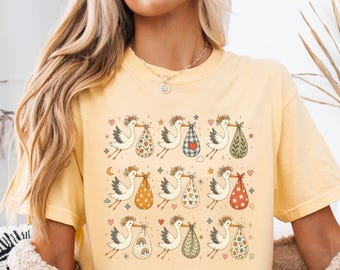 May include: A butter yellow t-shirt featuring a repeating grid pattern of storks carrying bundles. Each stork is depicted in a line-art style, with the bundles showcasing various patterns including florals, hearts, rainbows, and plaid designs.