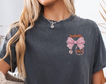 May include: A charcoal gray short-sleeve t-shirt featuring a graphic on the left chest. The design includes a pink patterned bow, a stethoscope, and the text Isabelle and NICU in orange lettering. The shirt has a crew neckline and a relaxed fit.