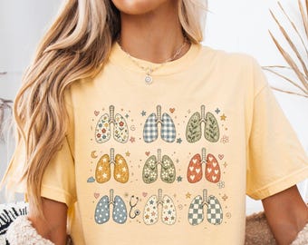 May include: A butter yellow t-shirt featuring a graphic print of nine pairs of lungs arranged in a grid. Each pair of lungs displays a different pattern, including floral, checkered, star, and heart designs, interspersed with small stars and hearts.