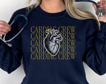 May include: A navy blue long-sleeve sweatshirt featuring a central graphic of a realistic anatomical heart. The text "CARDIAC CREW" is repeated in gold lettering behind the heart illustration. A stethoscope is draped around the neck area.