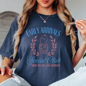 Op de afbeelding: Een marineblauw T-shirt met de tekst "Early Arrivals Social Club Where Due Dates Are Overrated" in roze en blauwe letters. Een roze en blauwe babypop-illustratie bevindt zich in het midden van de tekst.