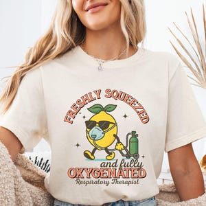 May include: Cream-colored t-shirt with a cartoon lemon wearing sunglasses and a face mask, holding an oxygen tank. The text reads "FRESHLY SQUEEZED and fully OXYGENATED Respiratory Therapist". The word "Natural" is printed at the bottom.