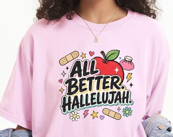 May include: A light pink t-shirt featuring a graphic print with the text "ALL BETTER HALLELUJAH" in black, a red apple, adhesive bandages, a pink ice pack, and small colorful flowers, stars, and hearts surrounding the central design.
