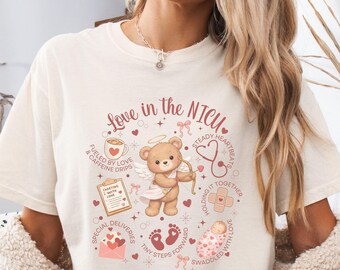 May include: Ivory-colored t-shirt with the text "Love in the NICU" and a graphic of a teddy bear angel with a bow and arrow. The design includes hearts, a coffee cup, a stethoscope, and baby footprints.