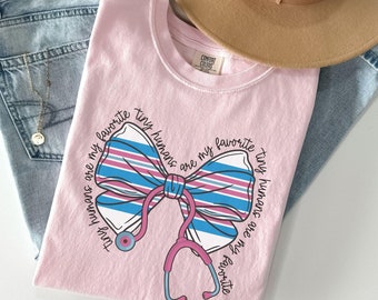 May include: Pink t-shirt with a stethoscope and a bow with the text "tiny humans are my favorite" repeated on both sides of the bow.