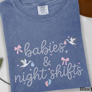 K&ouml;nnte beinhalten: Blaues Jeans-T-Shirt mit den Worten "babies & night shifts" in Schreibschrift. Das Design umfasst rosa Schleifen, St&ouml;rche, die Babys tragen, und Babyf&uuml;&szlig;e. Das Shirt ist aus einem weichen Material.