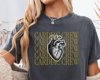 May include: A charcoal gray short-sleeve t-shirt featuring a repeating gold text pattern that reads CARDIAC CREW. Centered on the chest is a detailed black and white anatomical heart illustration with a white outline. The shirt is labeled as Pepper.