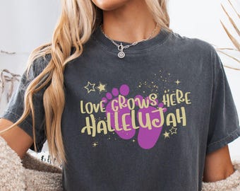 May include: A charcoal gray short-sleeve t-shirt featuring a graphic print with the text "LOVE GROWS HERE HALLELUJAH" in yellow, accompanied by purple baby footprints and small yellow star accents. The shirt has a crew neckline.
