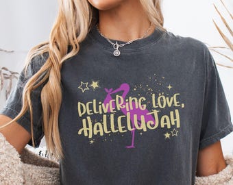 May include: A charcoal gray short-sleeve t-shirt featuring a graphic of a pink stork carrying a bundle. The text reads "DELIVERING LOVE, HALLELUJAH" in yellow lettering, accented by yellow stars and small dots. The shirt has a crew neckline and a relaxed fit.