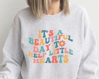 May include: A gray sweatshirt with the text "It's a beautiful day to help little hearts" in colorful, retro-style lettering. The text is surrounded by a flower and stars.