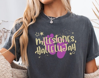 May include: A charcoal gray short-sleeve t-shirt featuring a graphic print with the yellow text "MILESTONES, HALLELUJAH" centered above two purple baby footprints. The design is accented with small yellow stars and sparkles on the front chest.