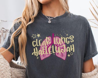 May include: A charcoal gray short-sleeve t-shirt featuring a graphic of pink lungs with the yellow text "CLEAR LUNGS HALLELUJAH" surrounded by small yellow stars and sparkles. The shirt has a crew neckline and a relaxed, vintage-washed appearance.