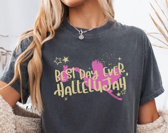 May include: A charcoal gray short-sleeve t-shirt featuring a pink stork silhouette carrying a bundle, with the yellow text "BEST DAY EVER, HALLELUJAH" and small yellow star accents printed on the front center of the garment.