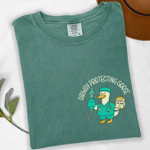 May include: Light green t-shirt with a crew neck. The shirt features a cartoon goose in a doctor's outfit holding a medical device and a clipboard. The text "AIRWAY PROTECTING GOOSE" is arched above the goose.