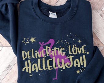 May include: A navy blue crewneck sweatshirt featuring a graphic of a purple stork carrying a bundle. The text "DELIVERING LOVE, HALLELUJAH" is printed in gold lettering across the center, accented by gold stars and small sparkling dots.