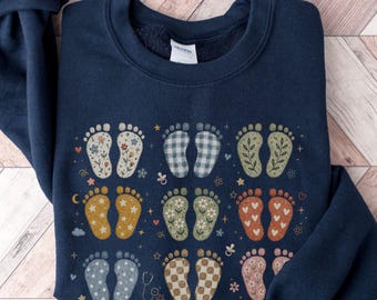 May include: A navy blue crewneck sweatshirt featuring a graphic print of nine pairs of baby footprints in various patterns, including floral, plaid, polka dot, and heart designs, interspersed with small stars, pacifiers, and a stethoscope icon.