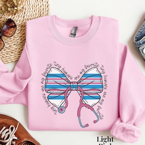 May include: Light pink sweatshirt with a graphic of a bow and stethoscope. The bow is blue and white striped. Text around the bow reads "Tiny humans are my favorite". The sweatshirt is a crew neck style.