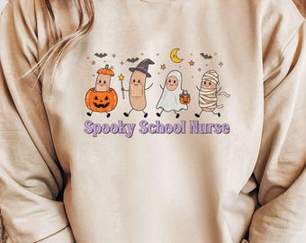 May include: A sand-colored crewneck sweatshirt featuring a graphic of four cartoon bandages dressed in Halloween costumes, including a pumpkin, a witch, a ghost, and a mummy. The text Spooky School Nurse is printed in purple below the characters.