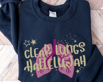 May include: A navy blue crewneck sweatshirt featuring a graphic of pink lungs with a dark pink bronchial tree. The text "CLEAR LUNGS HALLELUJAH" is printed in a light gold font across the center, surrounded by small gold stars and sparkles.