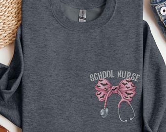 May include: A dark heather gray crewneck sweatshirt featuring a graphic on the left chest. The design includes the text "SCHOOL NURSE" in a light gray font above a pink bow patterned with small black bats, connected to a medical stethoscope.