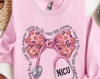 May include: A light pink crewneck sweatshirt featuring a large pink bow graphic patterned with small orange pumpkins and black baby footprints. A stethoscope is draped over the bow with the text NICU and Caring for the cutest little pumpkins.