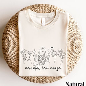 May include: A natural colored t-shirt with a black and white illustration of a baby swaddled in a blanket with flowers and butterflies. The text "neonatal icu nurse" is printed below the illustration.