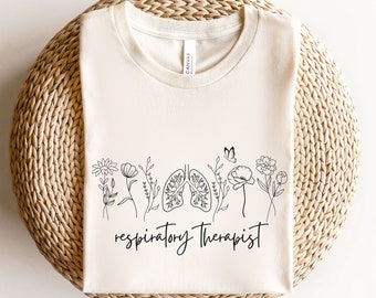 May include: A natural colored t-shirt with a black line drawing of lungs and flowers. The text "respiratory therapist" is written below the drawing.