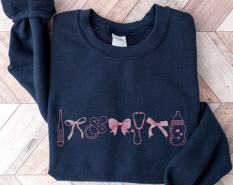 May include: A navy blue sweatshirt with a pink graphic of a thermometer, pacifier, bow, stethoscope, bow, and baby bottle.